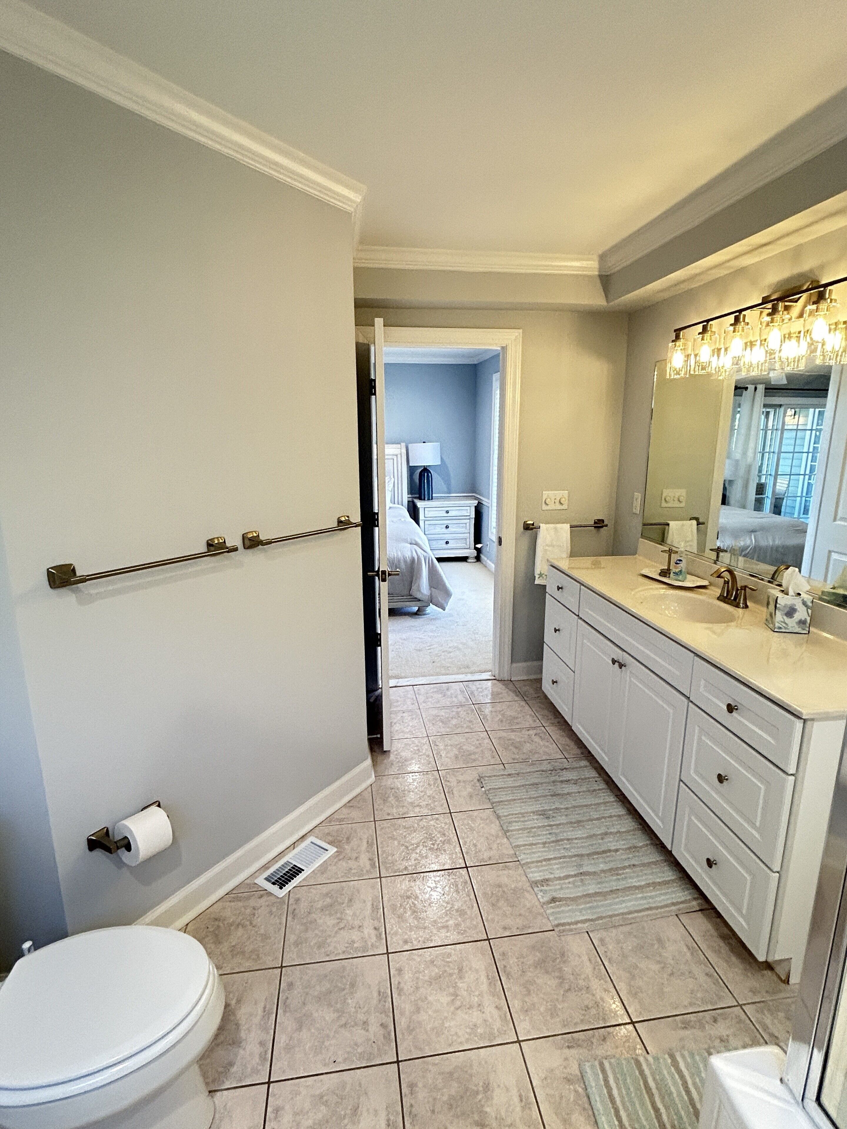 A bathroom with a toilet, a sink, and a mirror. There is a towel rack above the toilet and a rug on the floor.