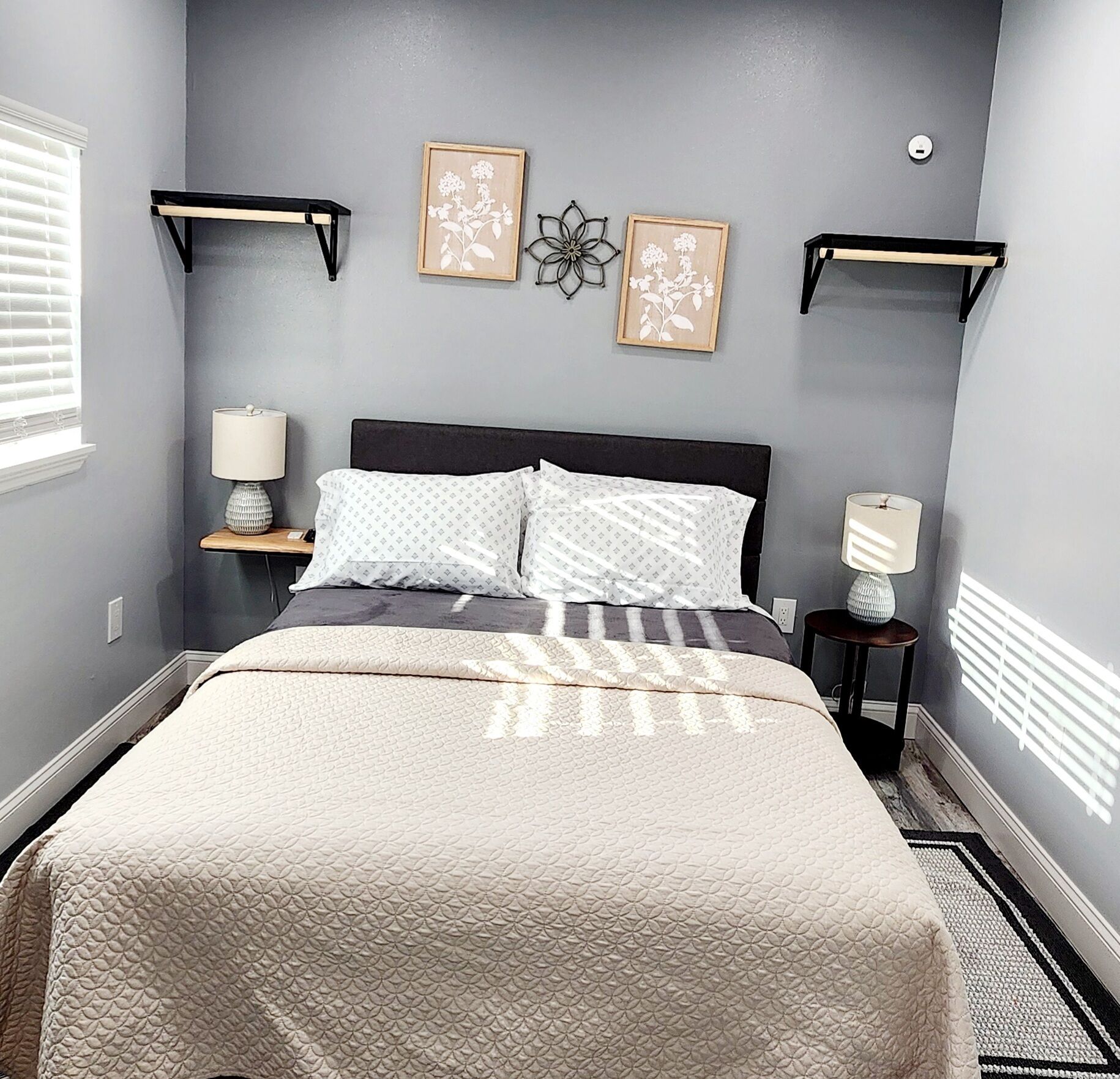 A bedroom with a bed, two bedside tables, a wall-mounted shelf, and a window with blinds.