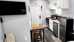Una cocina compacta con armarios blancos, una estufa, un fregadero y un refrigerador. Hay una mesa de madera y una silla en la esquina.