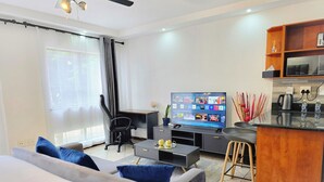 A modern living room with a sofa, a coffee table, a TV, a desk, and a kitchen area with a microwave and a kettle.