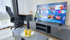 A modern living room with a flat-screen TV displaying a streaming service interface, a black office chair, a grey sofa, and a small table with decorative items.