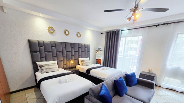 A hotel room with two beds, a grey sofa, a ceiling fan, and a large window with curtains.