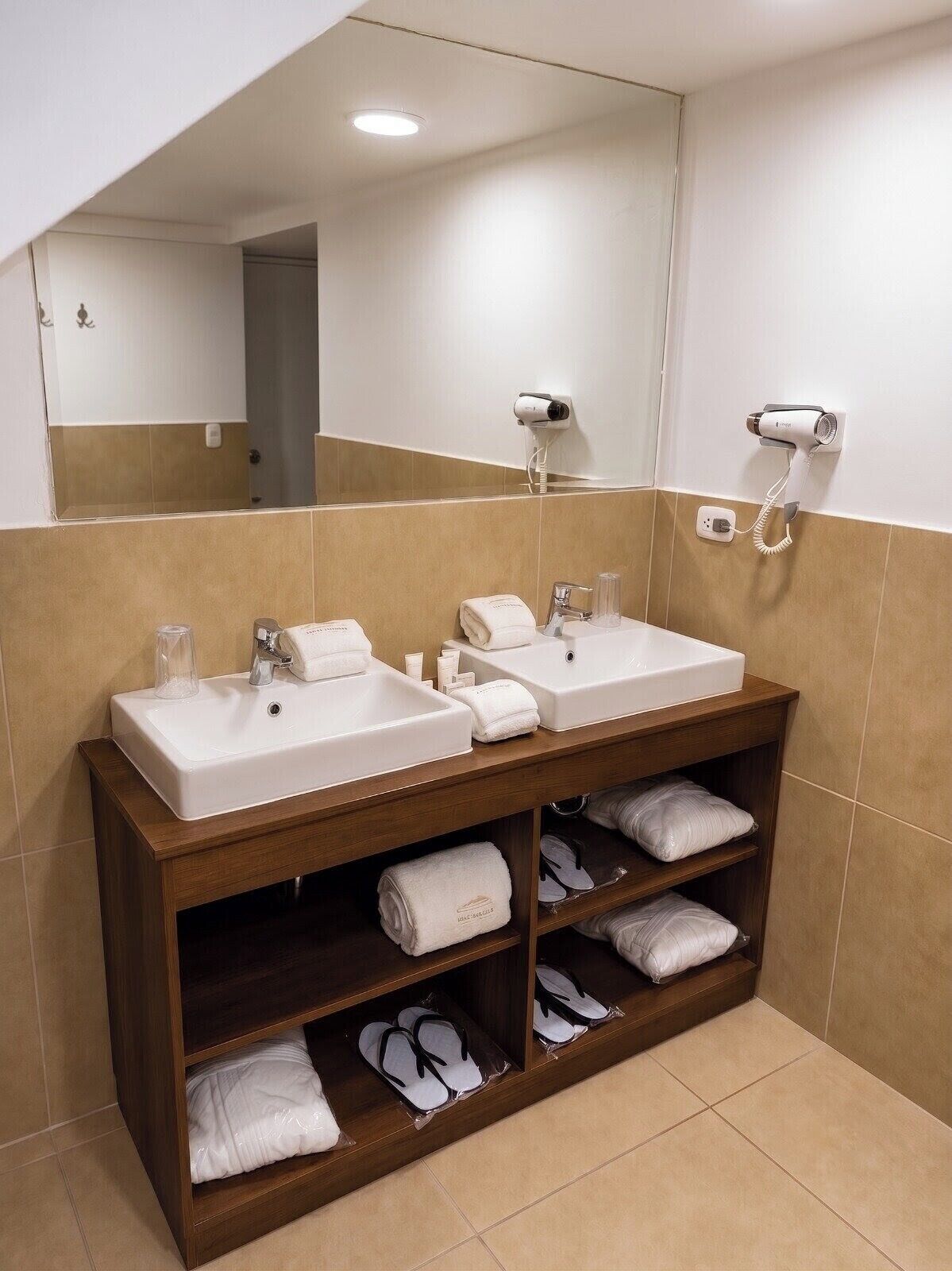 A bathroom with two sinks, a mirror, and a shelf with towels and slippers.