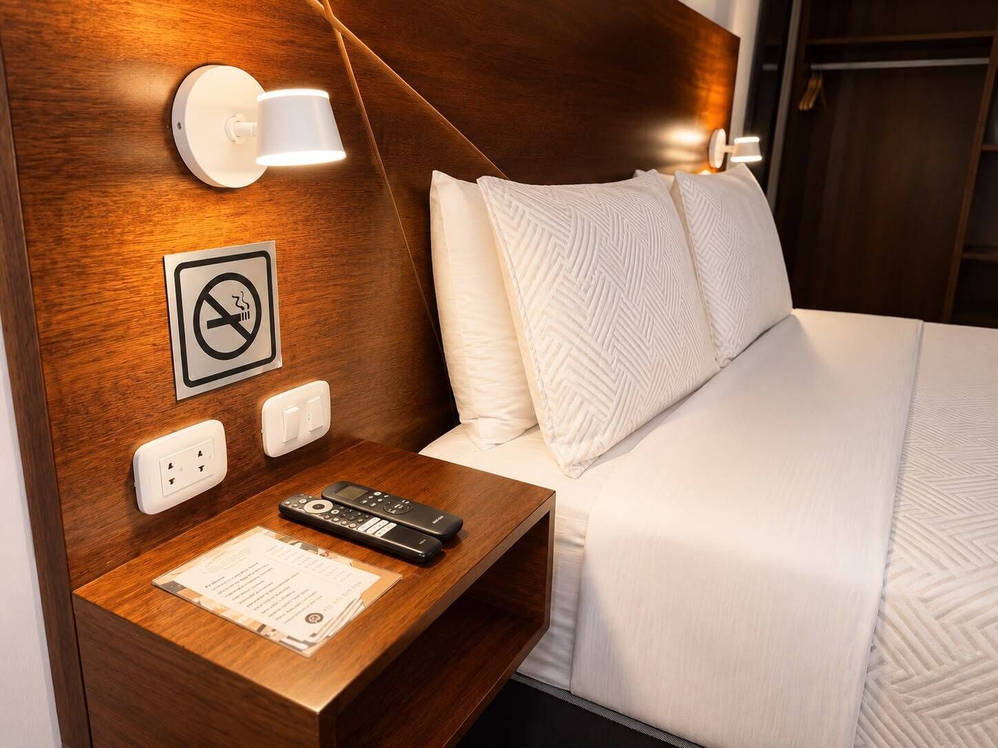 Habitación de hotel con cabecera de madera, mesita de noche con control remoto, cartel de no fumar y cama con ropa blanca.