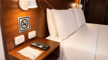 Habitación de hotel con cabecera de madera, mesita de noche con control remoto, cartel de no fumar y cama con ropa blanca.
