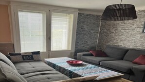 Een woonkamer met een grijze bank, een salontafel met een tafelkleed met patroon, en een grijze zithoek met rode kussens.