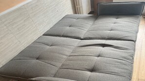 Een grijze bank met een hoge rugleuning en een beklede zitting, geplaatst tegen een houten vloer en een lichtgekleurde muur.