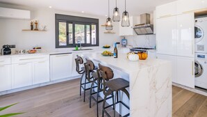 Une cuisine moderne avec des meubles blancs, un îlot en marbre, des tabourets de bar, un four encastré et un lave-linge.