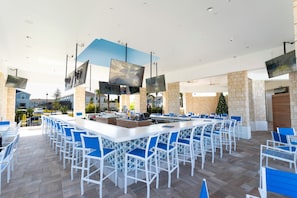 Un vaste espace extérieur aménagé pour les repas, avec des chaises blanches et bleues, un bar central et plusieurs téléviseurs à écran plat fixés au plafond.