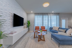A modern living room with a flat-screen TV, a blue sofa, two orange chairs, a coffee table, and a patterned rug.