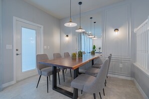 Comedor con mesa de madera, seis sillas grises, luces colgantes y una puerta blanca con ventanal.