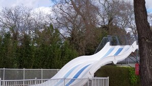 A white metal slide with blue stripes, a white fence, and a tree.