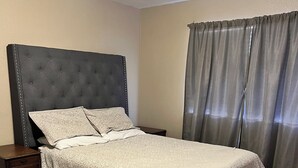 Ein Schlafzimmer mit einer gesteppten Kopfstütze, einem Bett mit gemusterten Betttextilien, einem Holz-Nachttisch und einem Fenster mit Vorhängen.