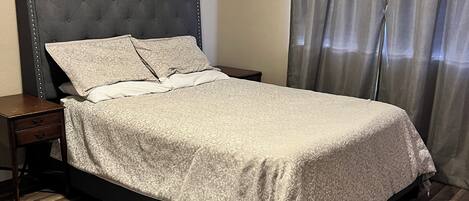 Een slaapkamer met een gestoffeerd hoofdeinde, een bed met beddengoed met een patroon, een houten nachtkastje en een raam met gordijnen.