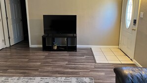 Ein Wohnzimmer mit Flachbildfernseher auf schwarzem Standfuß, Holzboden, einem gemusterten Teppich und einer Tür mit Fenster.