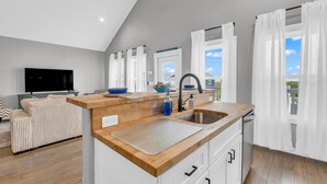 A modern kitchen with a wooden countertop, white cabinets, and a built-in dishwasher. There is a large window with white curtains, a television in the living area, and a sofa with striped cushions.