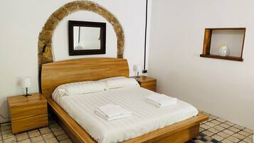 Una cama bien hecha con cabecera de madera, una mesita de noche con lámpara y un espejo montado en la pared.
