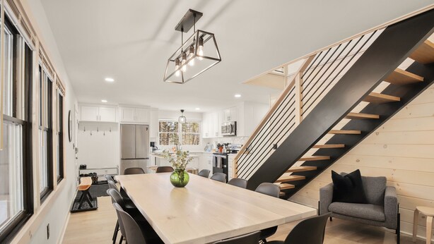 Una cocina moderna con armarios blancos, un comedor con mesa y sillas de madera, y una escalera con escalones de madera y barandillas de metal.