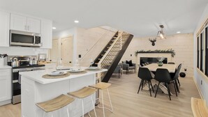 Una cocina moderna con armarios blancos, una isla central con taburetes, un comedor con mesa y sillas de madera, y una escalera con barandilla de madera.