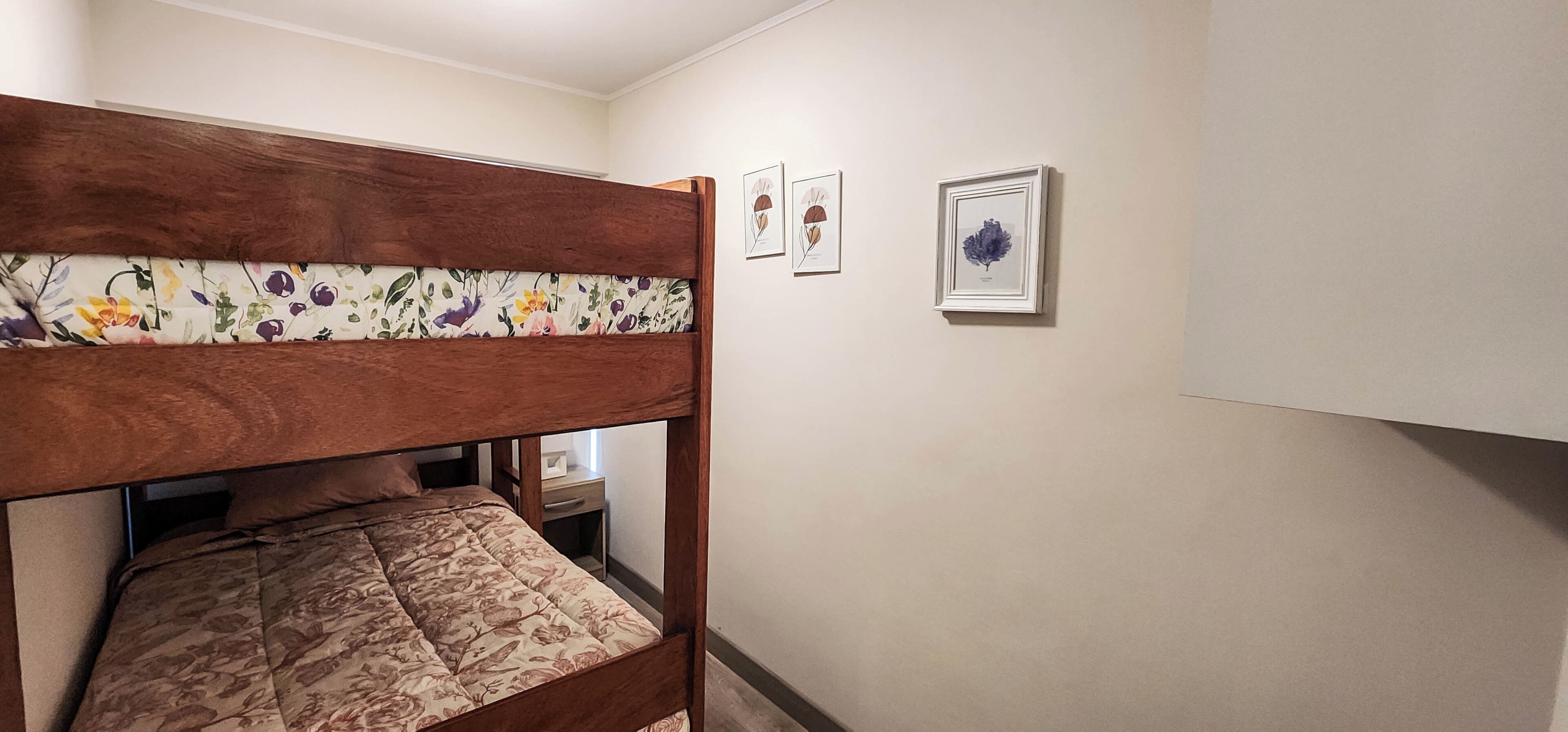 Una cama litera con ropa de cama floral, una mesita de noche y dos láminas botánicas enmarcadas en la pared.