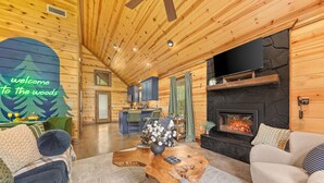 Ein gemütliches Blockhaus-Interieur mit Holzbalkendecke, Kamin aus Stein, Essbereich und Wohnzimmer mit großem Holztisch.