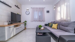 Un salon moderne avec un canapé gris, une table basse noire, un téléviseur posé sur un meuble en bois, une horloge au mur et une fenêtre ornée de rideaux légers.
