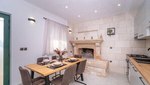 Une cuisine moderne avec une cheminée en pierre, un coin repas avec une table et des chaises en bois, et une vue sur l’extérieur par une porte.