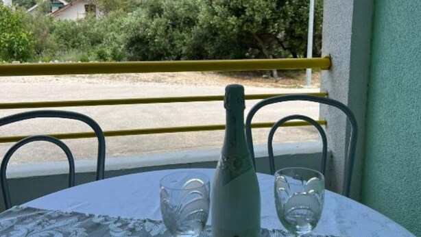 Une table avec une bouteille et deux verres, posée sur une nappe, d’où l’on aperçoit une zone côtière avec des maisons et de la végétation.