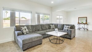 Amplia sala de estar con un sofá seccional gris, una mesa de centro blanca y ventanas grandes con vistas al exterior.