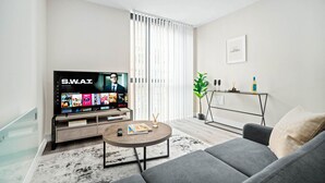 Ett modernt vardagsrum med en platt-TV som visar gränssnittet för en streamingtjänst, en grå soffa, ett soffbord med ett tidningsnummer och en minimalistisk inredning som inkluderar en växt och ett litet bord med en flaska.