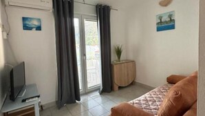 Habitació amb un sofà, una televisió sobre un suport, una petita taula auxiliar de fusta amb una planta i una porta que dona a l'exterior.