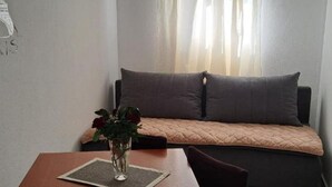 A small, cozy room with a wooden table, a vase with flowers, a cushioned bench, and a window with sheer curtains.