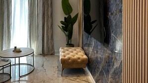 A modern living room with a marble floor, a tufted ottoman, a round glass coffee table, a large potted plant, and a flat-screen TV mounted on a marble wall. - Two Bedroom Apartment with Terrace and Swimming pool (Zagvozd)