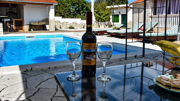 Ett bord vid poolkanten med en flaska kvassetino crno vino, två vinglas och en tallrik med mat.