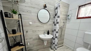 Ein modernes Badezimmer mit einem weißen Waschbecken, einem runden Spiegel, einer Dusche mit gemustertem Duschvorhang, einer Toilette und einem kleinen Ablagefach mit Pflegeprodukten.