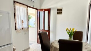 Ein kleiner Essbereich mit Tisch, bedeckt mit einem Tischläufer mit Punkten, zwei Stühlen und einer Vase mit gelben Blumen. Links steht ein weißer Kühlschrank, im Hintergrund eine Holztür mit Glasfenster.
