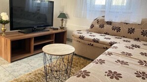 A living room with a flat-screen TV on a wooden stand, a round coffee table, a sofa with a floral pattern, and a window with sheer curtains. - One Bedroom Apartment with Balcony (3) (Biograd na Moru)