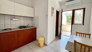 A compact kitchen with white cabinets, a sink, and a small stool. There is a dining area with a table and chairs, and an air conditioning unit above the window. - Apartments Fortuna - Studio Apartment (Biograd na Moru)