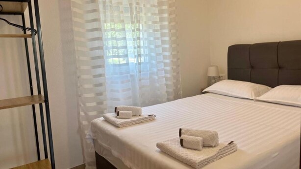 A neatly made bed with white linens and two towels, a bedside table with a lamp, and a window with sheer curtains. - Apartments Fortuna - Studio Apartment (Biograd na Moru)