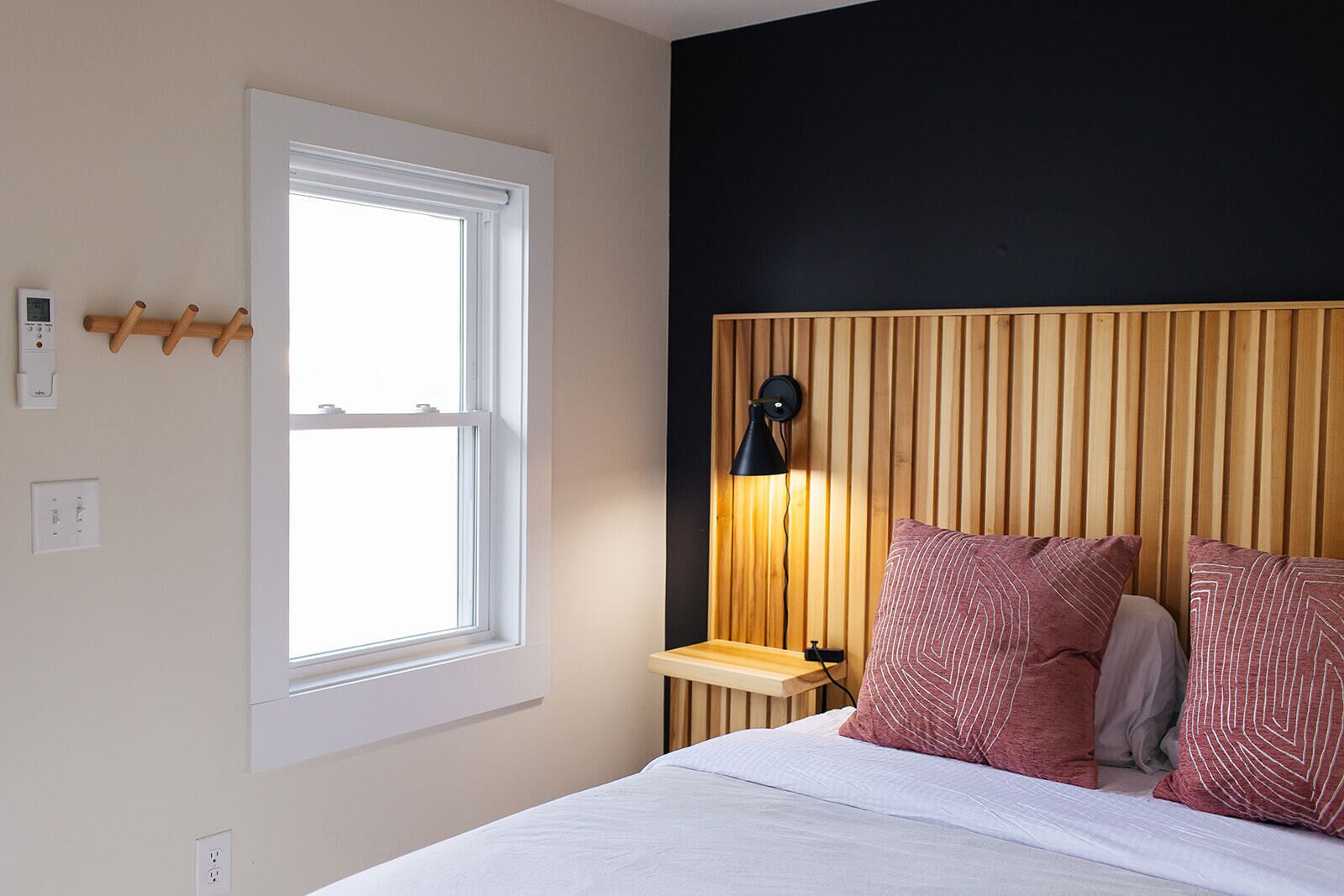 A bedroom with a bed, a bedside table, a lamp, a window, and a wall-mounted light fixture.