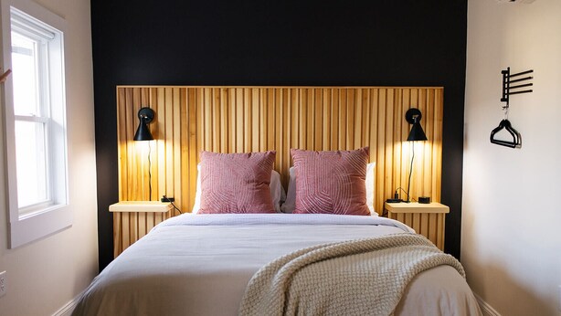 A bedroom with a wooden slat headboard, two bedside lamps, a bed with a white comforter and two pink pillows, a beige throw blanket, and a wall-mounted coat rack. - The Mane & Co - No. 4 (Chambersburg)