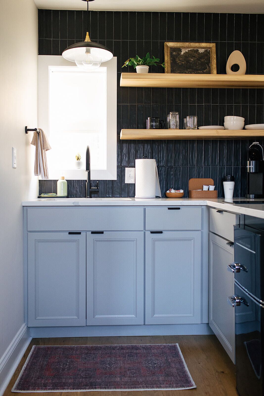 A kitchen with light blue cabinets, a wooden countertop, a hanging light fixture, a window, and a dark tiled backsplash.
