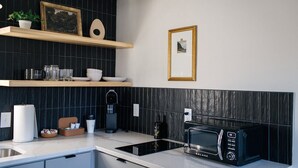 A modern kitchen with a black backsplash, white countertops, a built-in oven, a microwave, and a coffee maker. There are shelves with decorative items and a framed picture on the wall. - The Mane & Co - No. 4 (Chambersburg)
