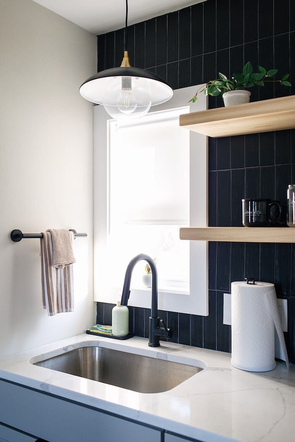 A modern kitchen with a stainless steel sink, a hanging pendant light, a towel rack, a window, and a shelf with a plant and a roll of paper towels.