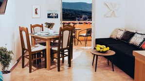 A dining area with a wooden table and chairs, a black sofa, and a small table with fruit. A balcony with a view of mountains is visible. - Hvar spa & apartments-1 Bedroom-Terrace/Sea View 3 (Vrboska)