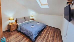 Ein Schlafzimmer mit geneigter Decke, einem Oberlicht, einem Bett mit gemustertem Überwurf, zwei Nachttischlampen und einem angebrachten Fernseher.