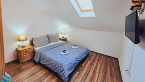 A bedroom with a sloped ceiling, a bed with a patterned duvet, two bedside lamps, a TV mounted on the wall, and a skylight. - One Bedroom Apartment with Bacony and Sea View (Vrboska)