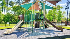 Ein Spielplatz mit Rutschen und Klettergerüsten, umgeben von einem Zaun und Bäumen.