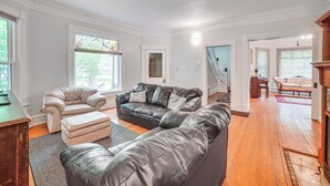 A spacious living room with a leather sofa, a matching ottoman, and a wooden coffee table. There is a staircase leading to an upper level, and a vintage armchair in the background. - Charming Champaign Victorian w/ Deck! (Champaign)