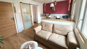Un salon moderne avec un canapé beige, un plancher en bois, une porte blanche et un coin cuisine avec un mur rouge et des plantes suspendues.
